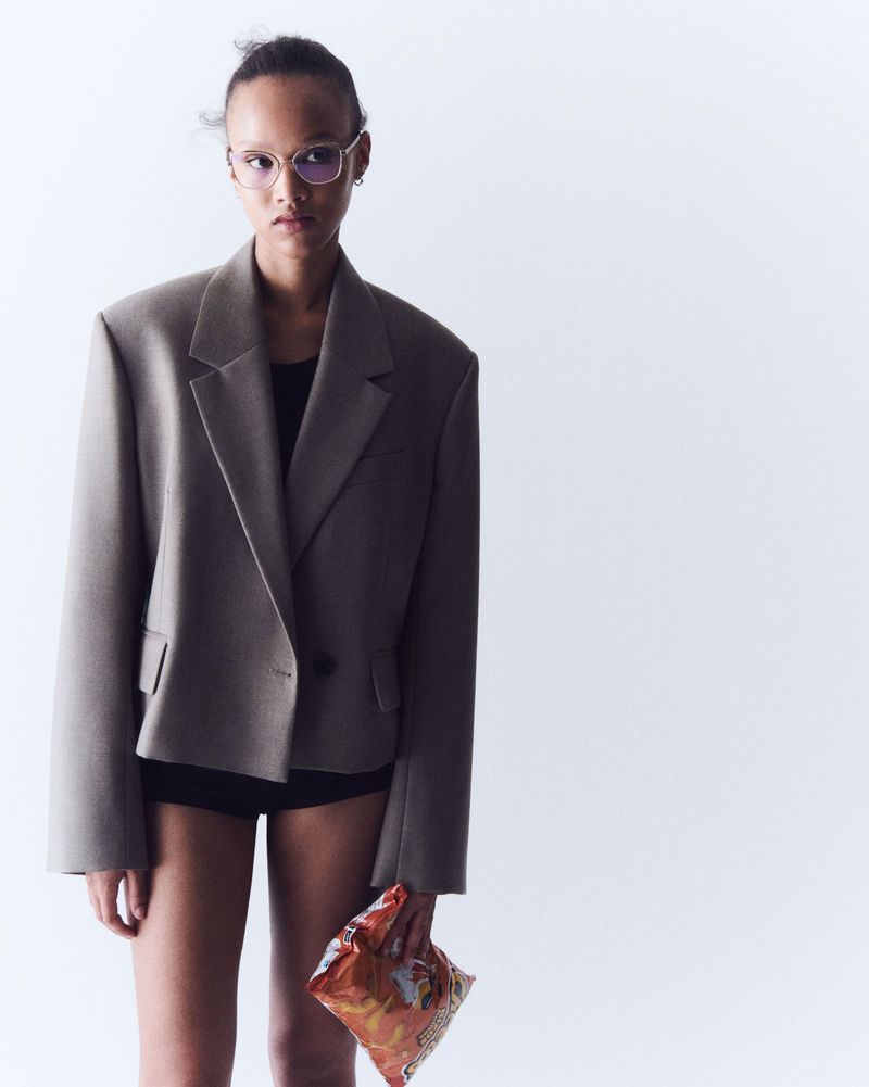 A fashion portrait of a woman in an oversized blazer and shorts posing against a white background, holding a bag of chips.