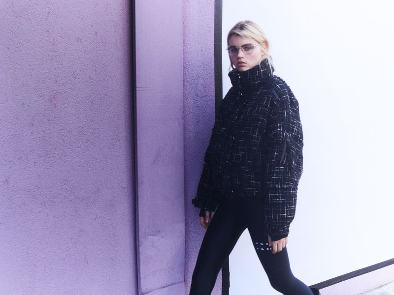 Adult female wearing a black jacket and black leggings posing in front of a lavender and white wall in an urban environment.