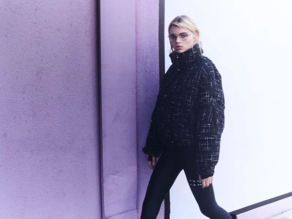 Model wearing a black tweed high-collar puffer jacket, glasses, and black leggings standing against a purple textured wall.