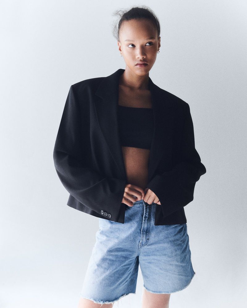 Portrait of a young adult woman with fair skin wearing a black jacket and denim shorts against a white studio backdrop. 
