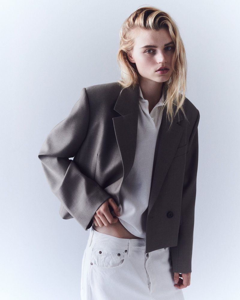 Fashion portrait of a blond woman in a grey blazer and white top and jeans posing indoors in a photo studio with soft lighting.