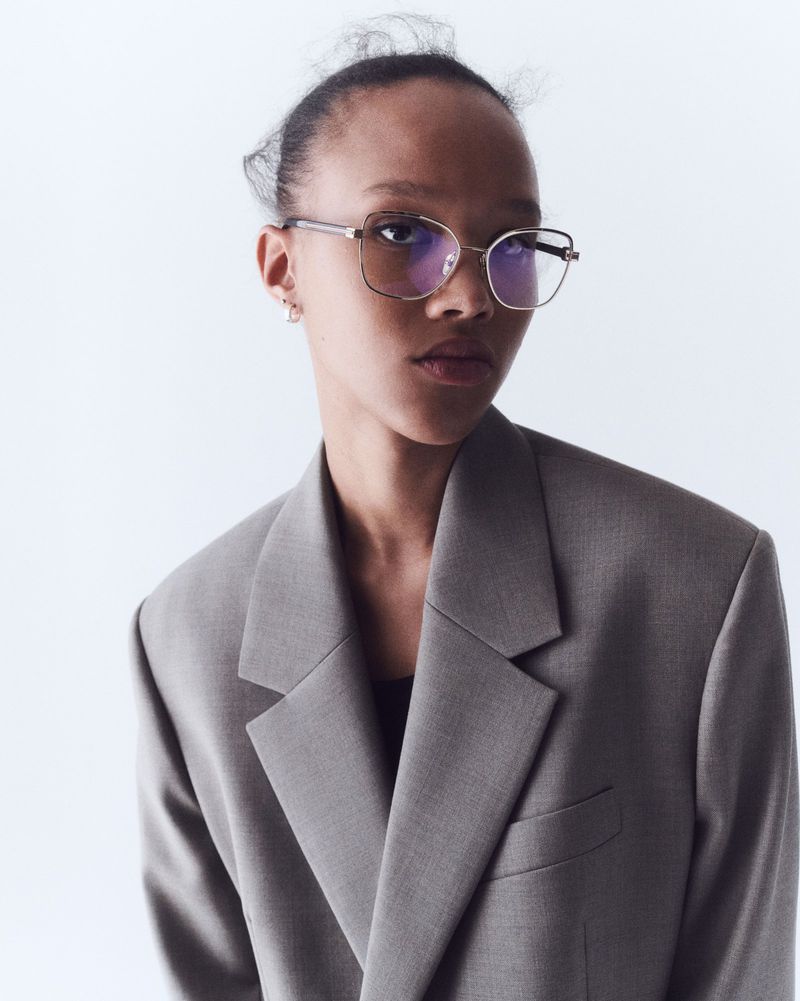 Woman in grey blazer and glasses poses against white background in a fashion shot with studio lighting and a neutral expression.