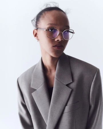 Woman in grey blazer and glasses poses against white background in a fashion shot with studio lighting and a neutral expression.