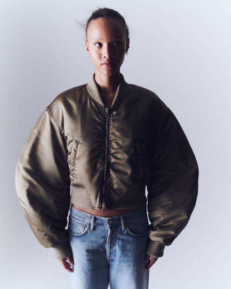 Woman in olive bomber jacket and jeans poses against a white backdrop in a fashion shot with diffused, even lighting and high-key tones.