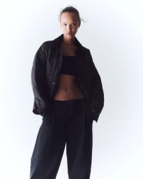 Model wearing an open black quilted jacket, black crop top, and wide-leg black trousers against a bright white studio backdrop.