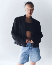 Model wearing an oversized black blazer and light wash denim shorts against a plain white studio wall