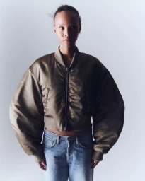 Model wearing an olive green cropped bomber jacket with voluminous sleeves and light wash jeans against a white studio background.
