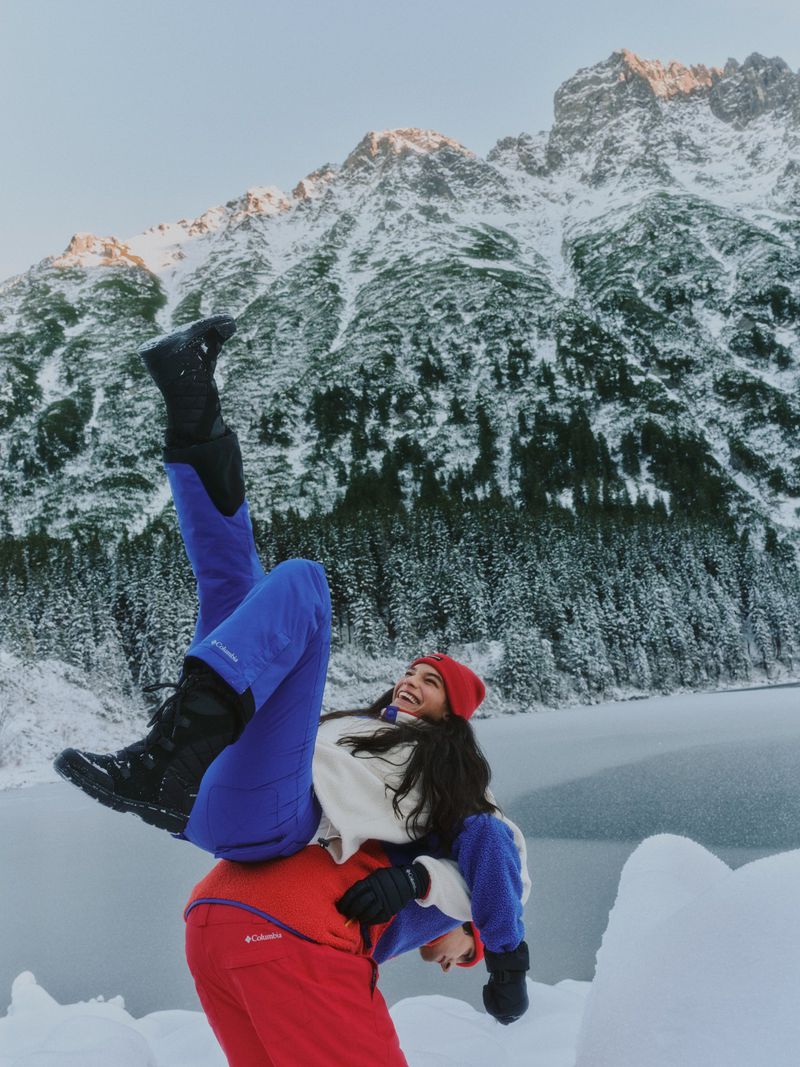 Woman in blue ski pants doing a handstand on a person wearing red ski pants in a snowy mountain landscape, backlit by golden light.