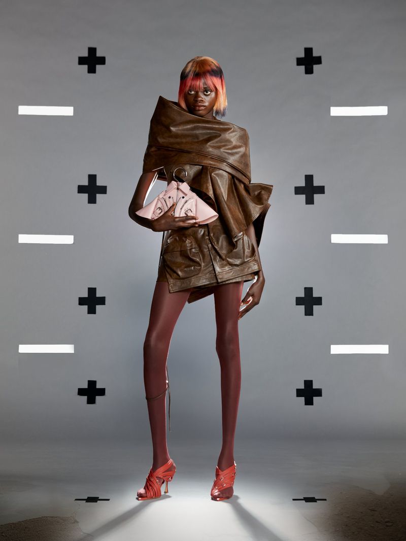 A model poses in a brown leather-like outfit and pink heels in a studio with geometric plus and minus signs on a gray backdrop.