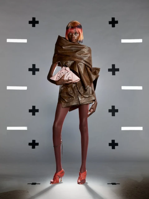 A model poses in a brown leather-like outfit and pink heels in a studio with geometric plus and minus signs on a gray backdrop.