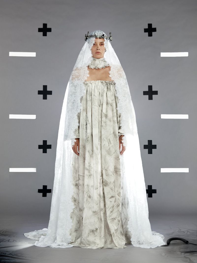 A lone woman in a long patterned gown and decorative crown stands centered against a stark gray background marked with black shapes.