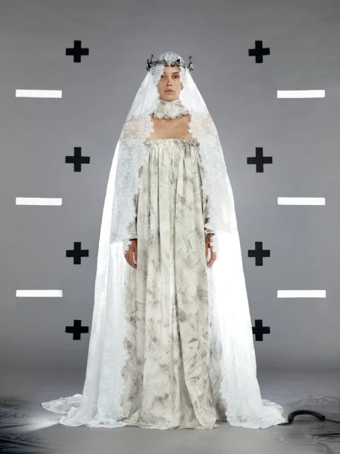 A lone woman in a long patterned gown and decorative crown stands centered against a stark gray background marked with black shapes.
