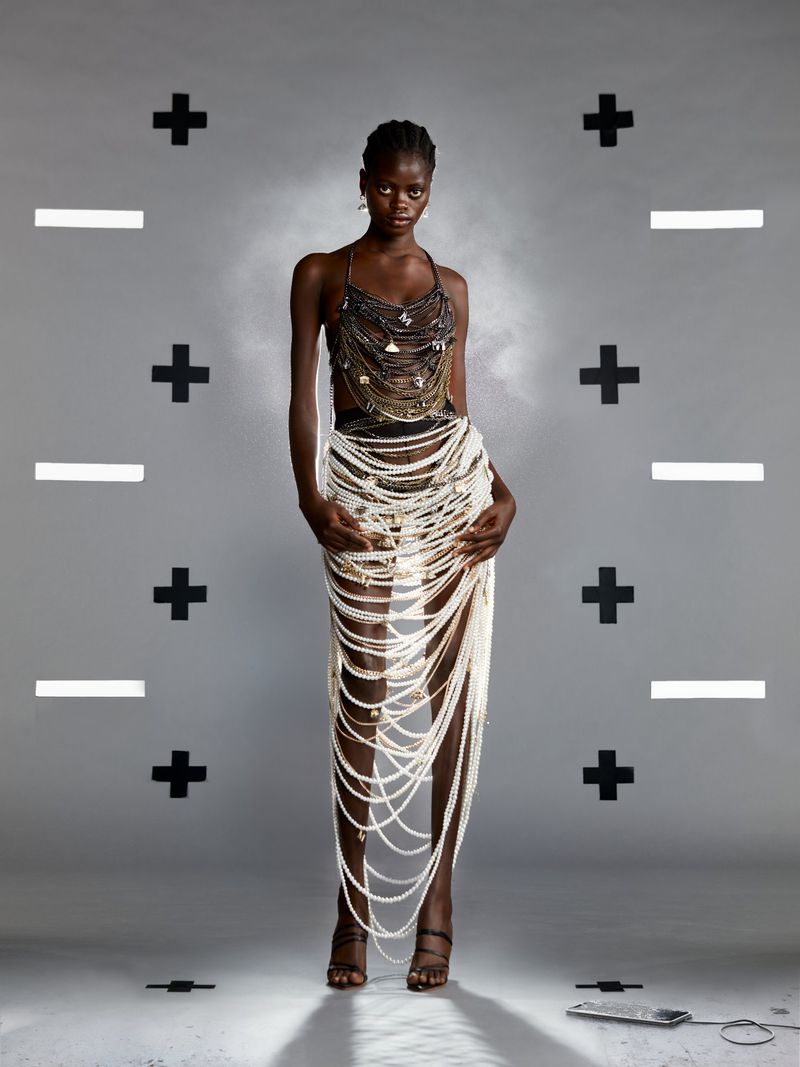 Full-length shot of an adult woman wearing a pearl skirt and beaded top, standing in front of a gray background with graphic plus and minus signs.