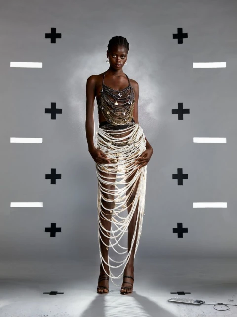 Full-length shot of an adult woman wearing a pearl skirt and beaded top, standing in front of a gray background with graphic plus and minus signs.