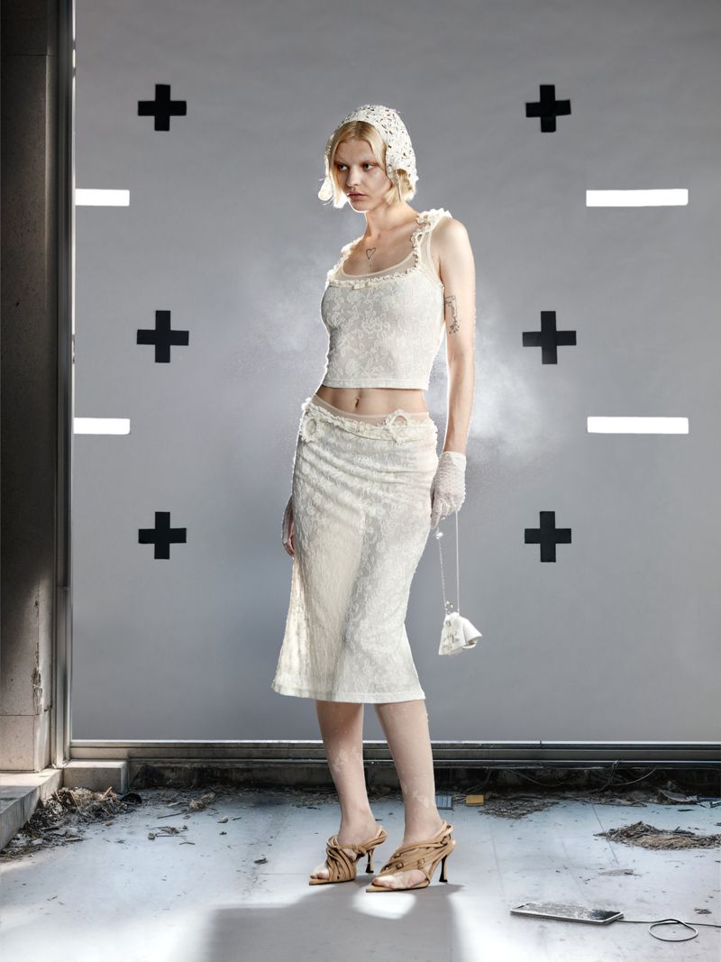 Young woman stands in a lace top, skirt, and headdress in a studio with plus and minus signs, holding a beaded purse and wearing strappy heels.