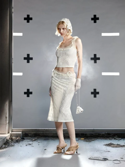 Young woman stands in a lace top, skirt, and headdress in a studio with plus and minus signs, holding a beaded purse and wearing strappy heels.