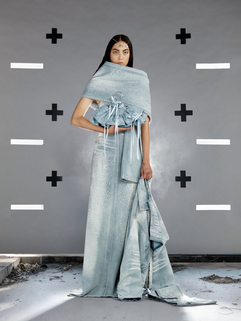 Fashion shot of a woman wearing a light blue gown and draped wrap standing against a patterned gray wall in a studio setting.