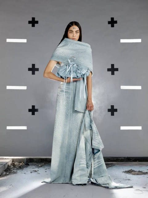 Fashion shot of a woman wearing a light blue gown and draped wrap standing against a patterned gray wall in a studio setting.