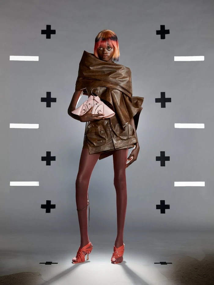 Model with dyed hair wearing brown structured wrap garment and pink handbag against a grey seamless background