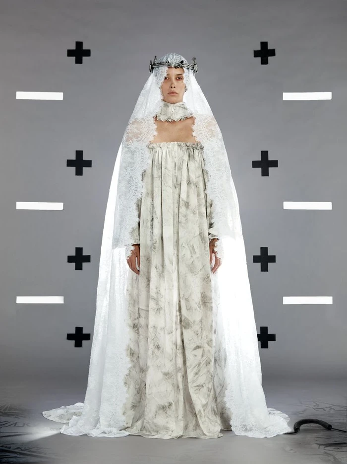 Model wearing a floor-length patterned white gown with a dramatic lace bridal veil and crown against a grey studio backdrop.