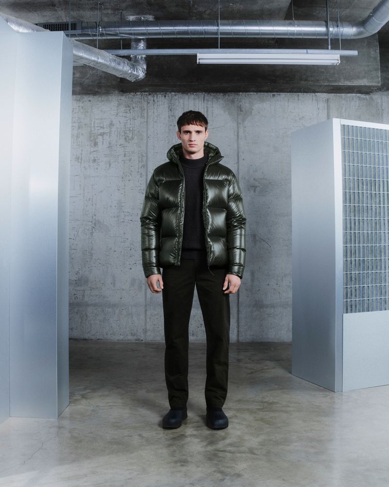 Full-length shot of a man in a green puffer jacket, black pants, and shoes, standing indoors against a gray concrete wall and metallic structures.