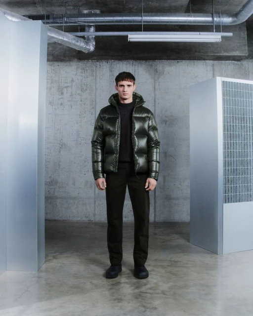 Full-length shot of a man in a green puffer jacket, black pants, and shoes, standing indoors against a gray concrete wall and metallic structures.