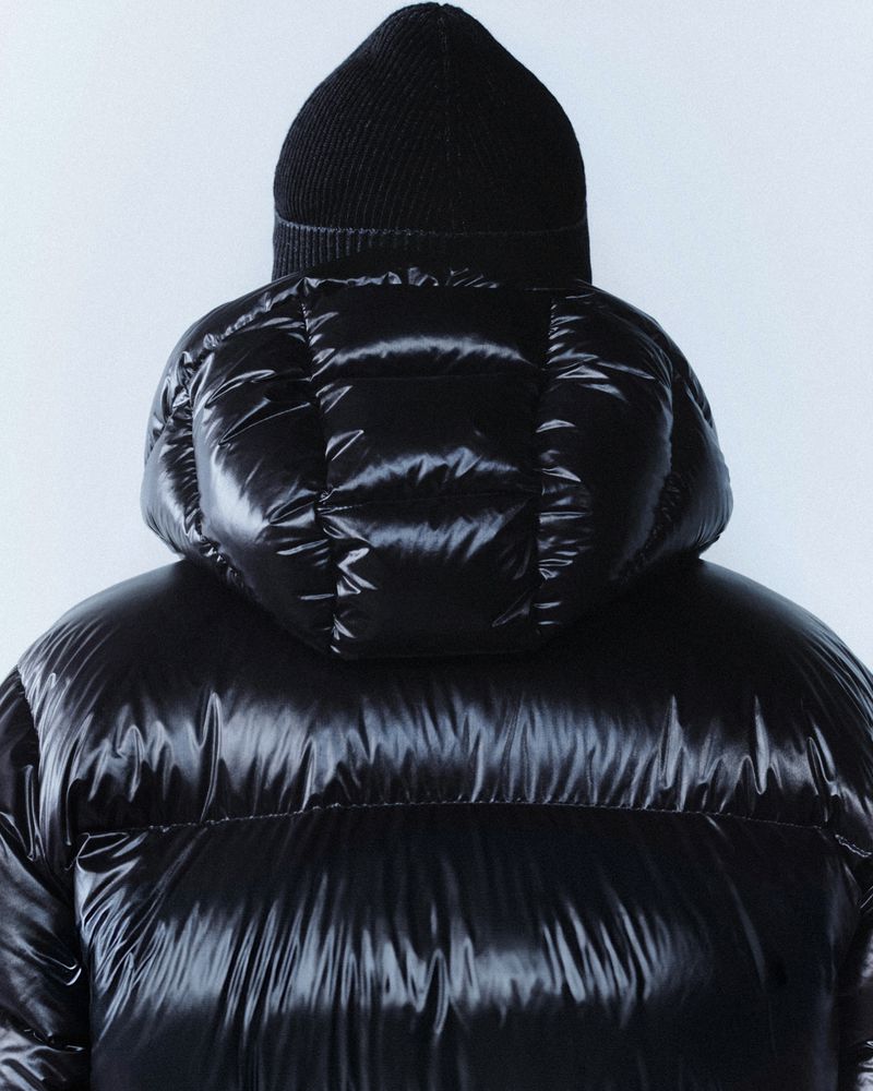 Close-up of a person from the back wearing a black puffer jacket with a hood and a black knit hat against a white background.