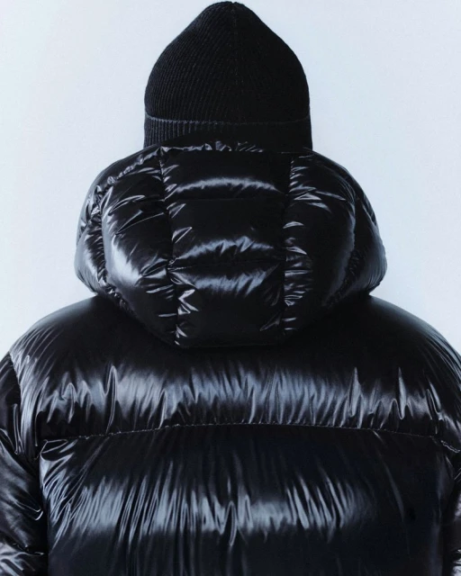 Close-up of a person from the back wearing a black puffer jacket with a hood and a black knit hat against a white background.