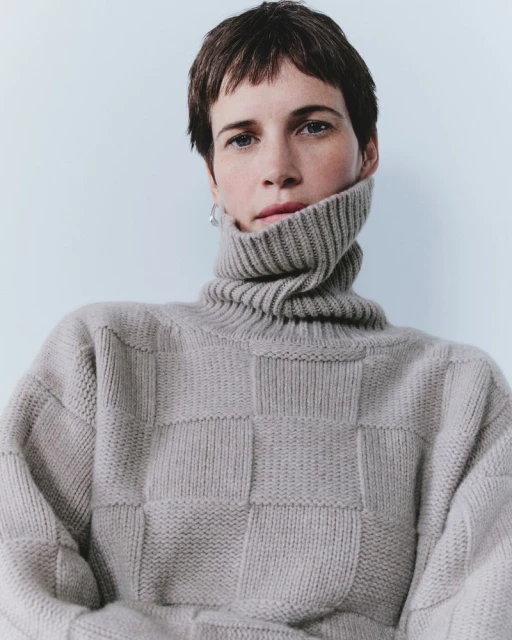 Close-up of a stylish woman with short hair wearing a checker-patterned turtleneck sweater against a pale blue background in a portrait.