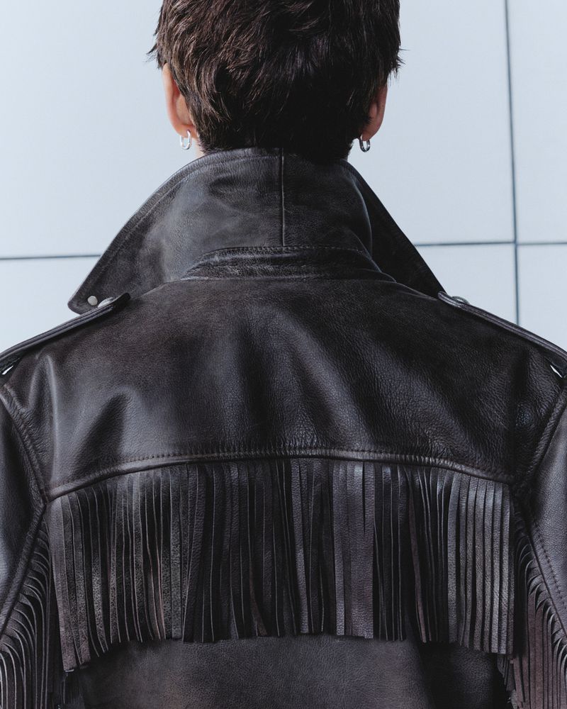 Back view of a person wearing a black leather jacket with fringe detailing against a light blue and white tile background.