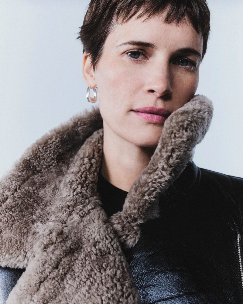 Close-up of a female fashion model in a leather jacket with a faux fur collar, posing in a photo studio with soft lighting.