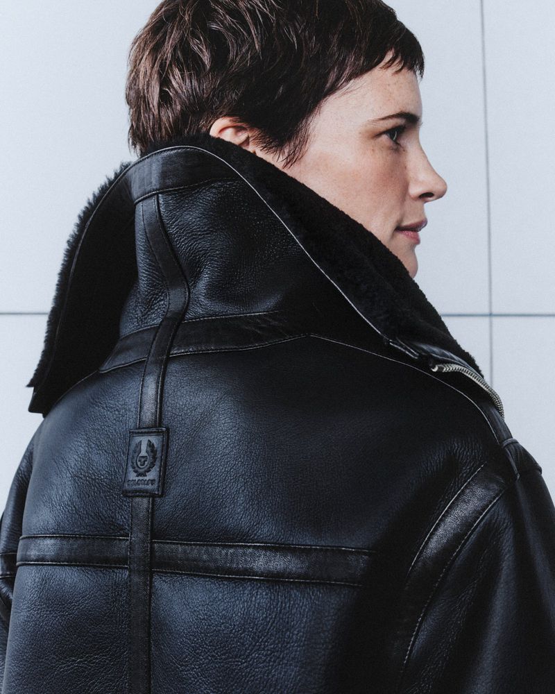 Profile shot of an adult female with short brown hair wearing a black leather aviator jacket against a white tiled background.