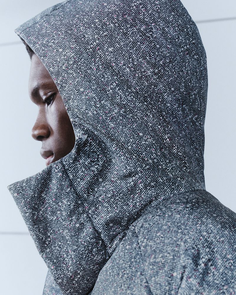 Close-up profile of a black man wearing a speckled grey hooded jacket with a high collar against a neutral white wall.