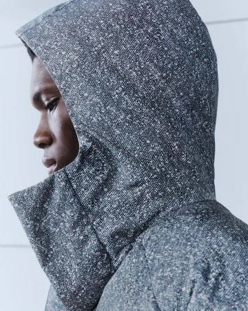 Close-up profile of a black man wearing a speckled grey hooded jacket with a high collar against a neutral white wall.