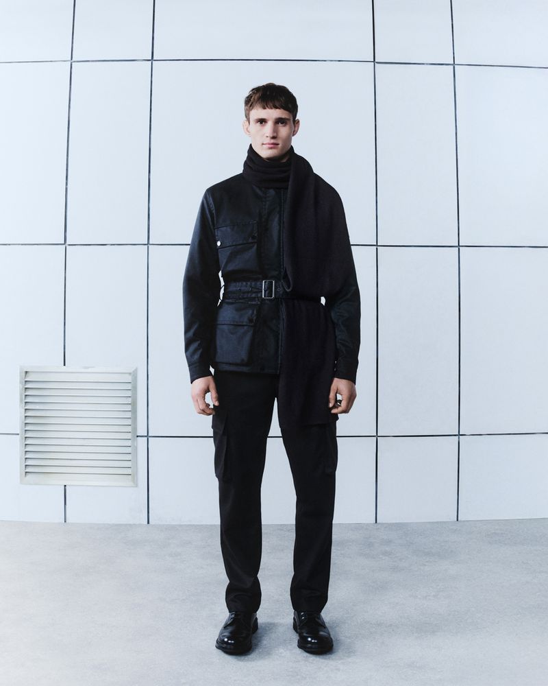 Full-length studio shot of a man in a black outfit with a jacket, pants, and scarf, posing against a white tiled background.