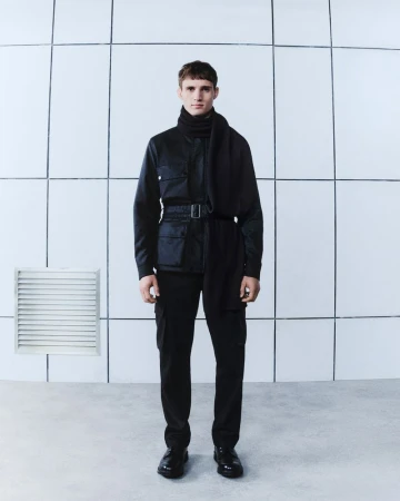 Full-length studio shot of a man in a black outfit with a jacket, pants, and scarf, posing against a white tiled background.