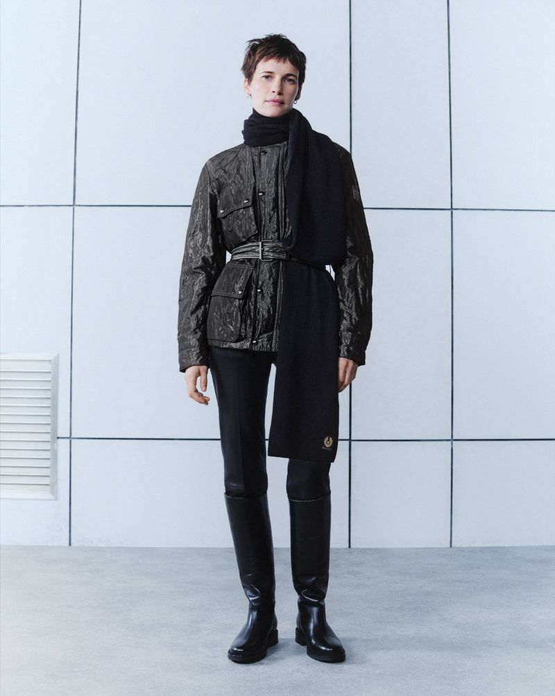 Full-length fashion shot of a woman with short brown hair wearing a belted black jacket, trousers, boots, and scarf against a white wall.