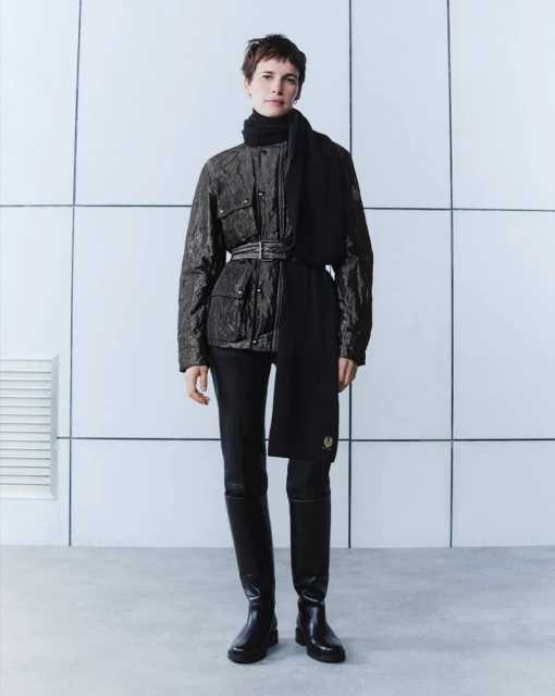 Full-length fashion shot of a woman with short brown hair wearing a belted black jacket, trousers, boots, and scarf against a white wall.
