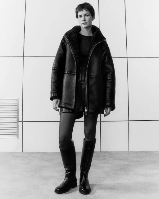 Monochrome fashion portrait of a woman in a shearling coat, black top, tights, mini skirt, and tall boots, posing against a tiled wall.