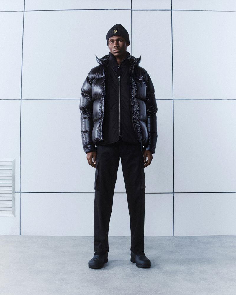 A Black male model in a black beanie, puffer jacket, and pants, stands in a modern studio with a minimalist, tiled white background.
