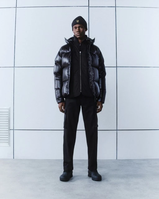 A Black male model in a black beanie, puffer jacket, and pants, stands in a modern studio with a minimalist, tiled white background.