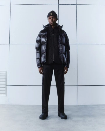 A Black male model in a black beanie, puffer jacket, and pants, stands in a modern studio with a minimalist, tiled white background.
