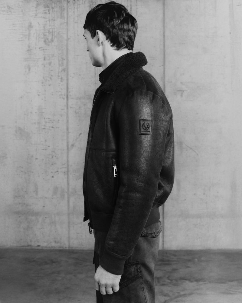 Monochrome fashion portrait of an adult male in a black shearling bomber jacket in front of a textured wall in a studio setting.