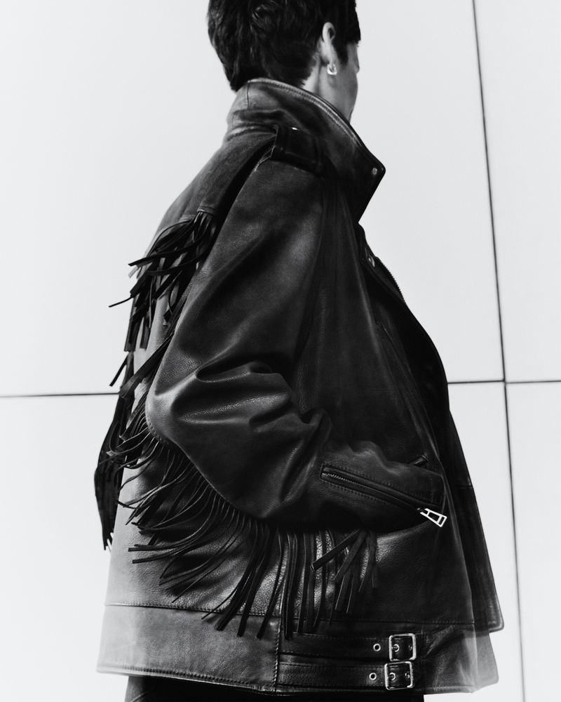 Back view of person wearing a black fringe leather jacket. Fashion shot. The person is positioned against a bright white background.