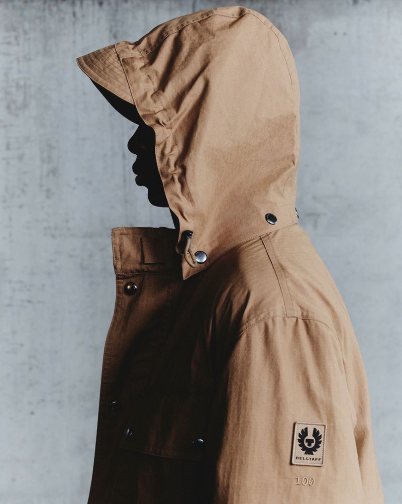 Tan hooded jacket detail on model showcasing fashion brand logo. The jacket is against a textured grey studio backdrop.