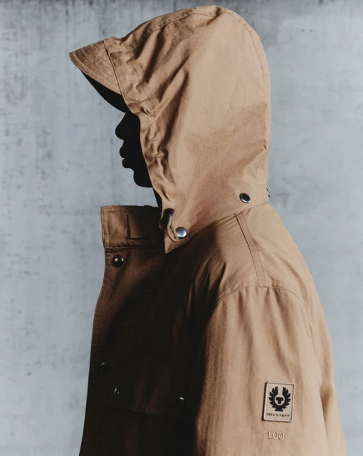 Tan hooded jacket detail on model showcasing fashion brand logo. The jacket is against a textured grey studio backdrop.