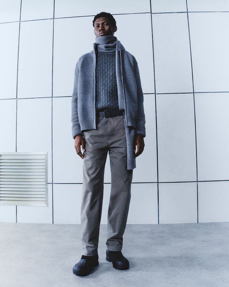 Full-length shot of an African American man in gray turtleneck, gray jacket, and gray trousers against a geometric white backdrop.