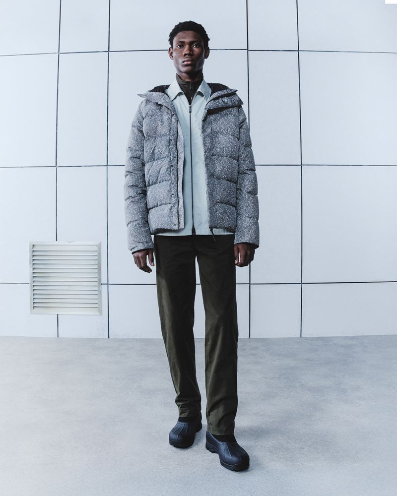 A dark-skinned male model is posing in a light gray puffer jacket, a light green collared shirt, dark green pants and black rubber shoes in a studio.