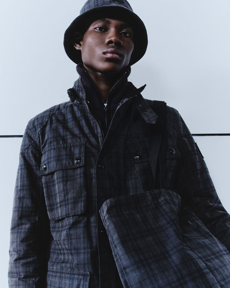 Dark-skinned male model in patterned jacket and bucket hat poses against a white wall, showcasing fashion in a studio setting.