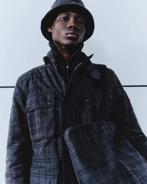 Dark-skinned male model in patterned jacket and bucket hat poses against a white wall, showcasing fashion in a studio setting.